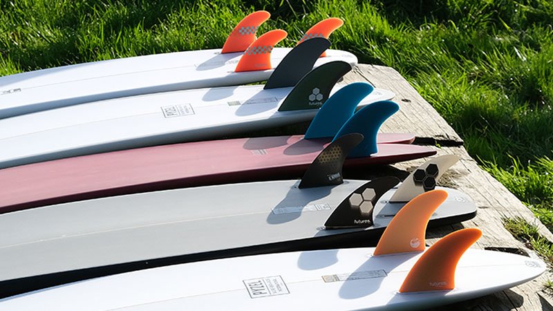 Conjunto de tablas de surf alineadas en una fila sobre un borde de madera, con aletas de colores naranja, negro, azul y gris, sobre césped verde. Conjunto de tablas de surf alineadas en una fila sobre un borde de madera, con aletas de colores naranja, negro, azul y gris, sobre césped verde.