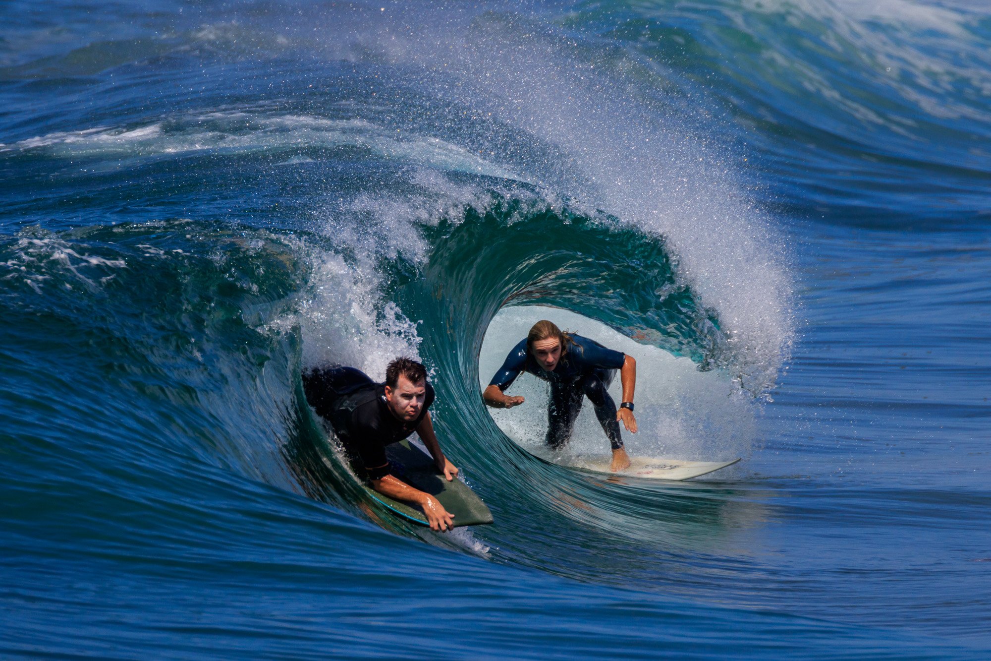 Dos surfistas montan una ola verde dentro del tubo, con agua azul alrededor y spray blanco. Incluye el keyword si procede, pero no se proporcionó en contexto. Alt text: Dos surfistas dentro de un túnel de ola, realizando maniobra enmarcada por el agua azul y el estallido de spray.