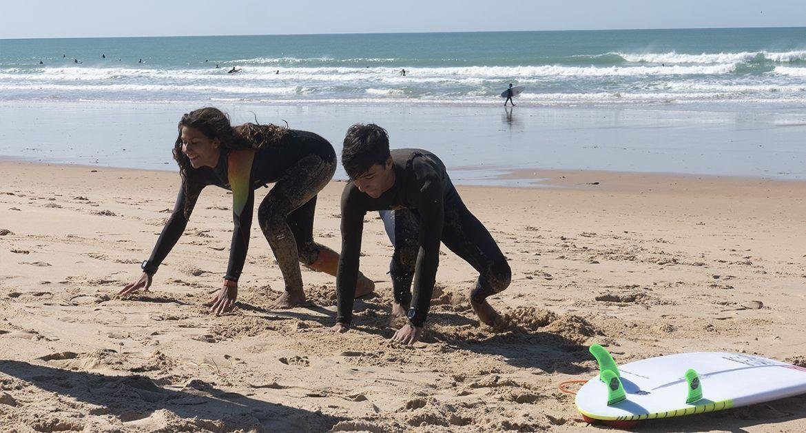 Due surfisti in muta si abbassano sulla sabbia pronta per partire, con tavola da wind surf sulla spiaggia e onde sullo sfondo.