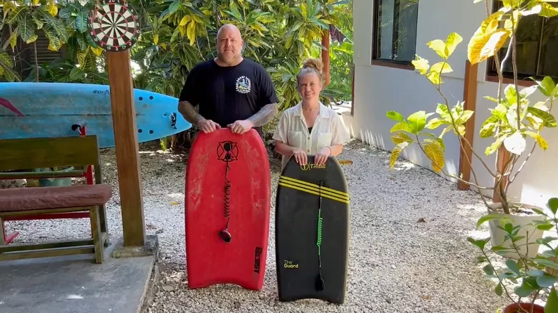 Dupla segurando pranchas de bodyboard em área externa com plantas; ao fundo itens de prancha e brinquedos. Se vê red bodyboard e prancha verde-escura ao lado.