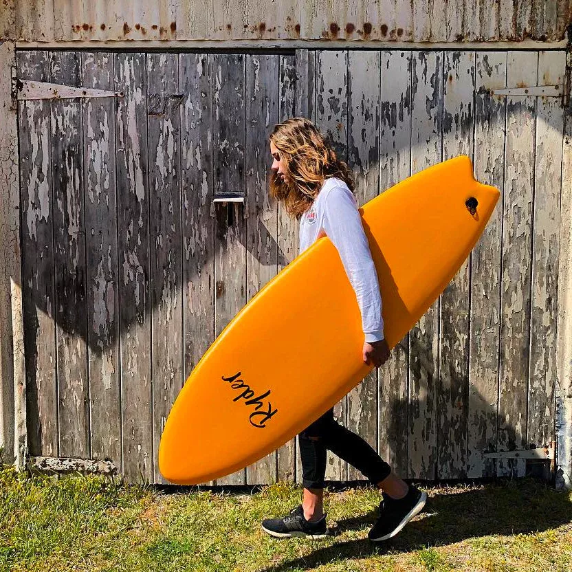 Estudiante camina junto a una cerca de madera sosteniendo una tabla de surf naranja con el logo “Kapow” sobre la camiseta blanca.