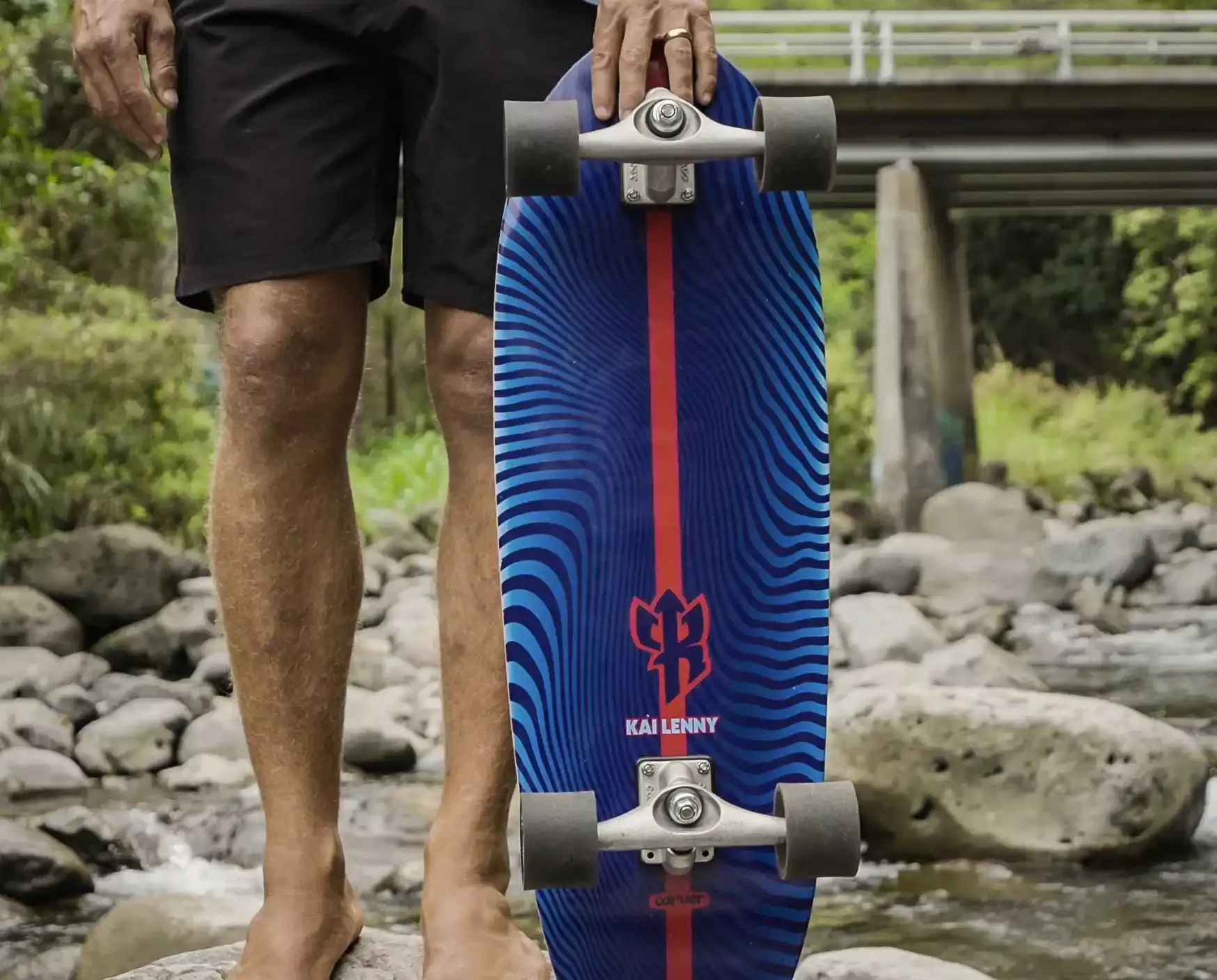 Longboard in primo piano, mani e piedi dell’utente vicino a una tavola colorata con ruote, in ambiente naturale vicino a un corso d’acqua.