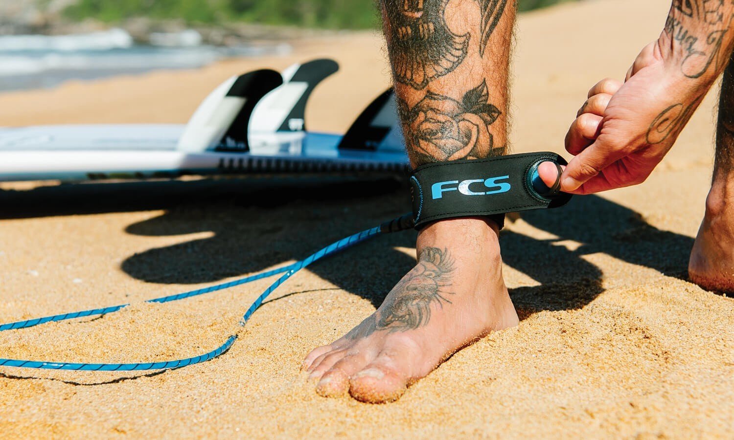 Polso con cavigliera di tipo leash FKIS durante prep per surf sulla sabbia, attaccata a tavola da surf in spiaggia.