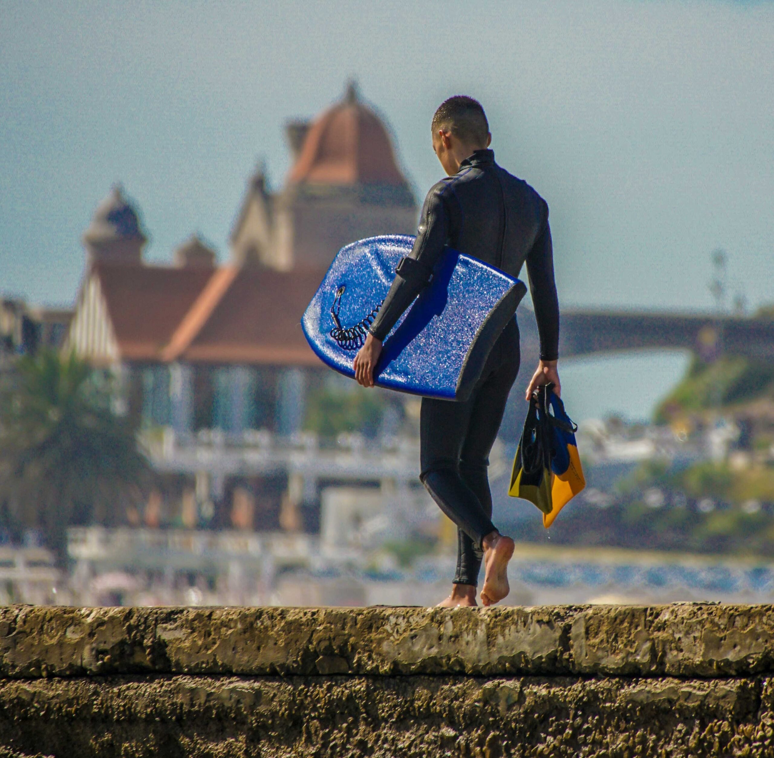 Prancha de surf colorida azul, com traje de neoprene, caminhando com bagagem à beira-mar, cenário urbano ao fundo.