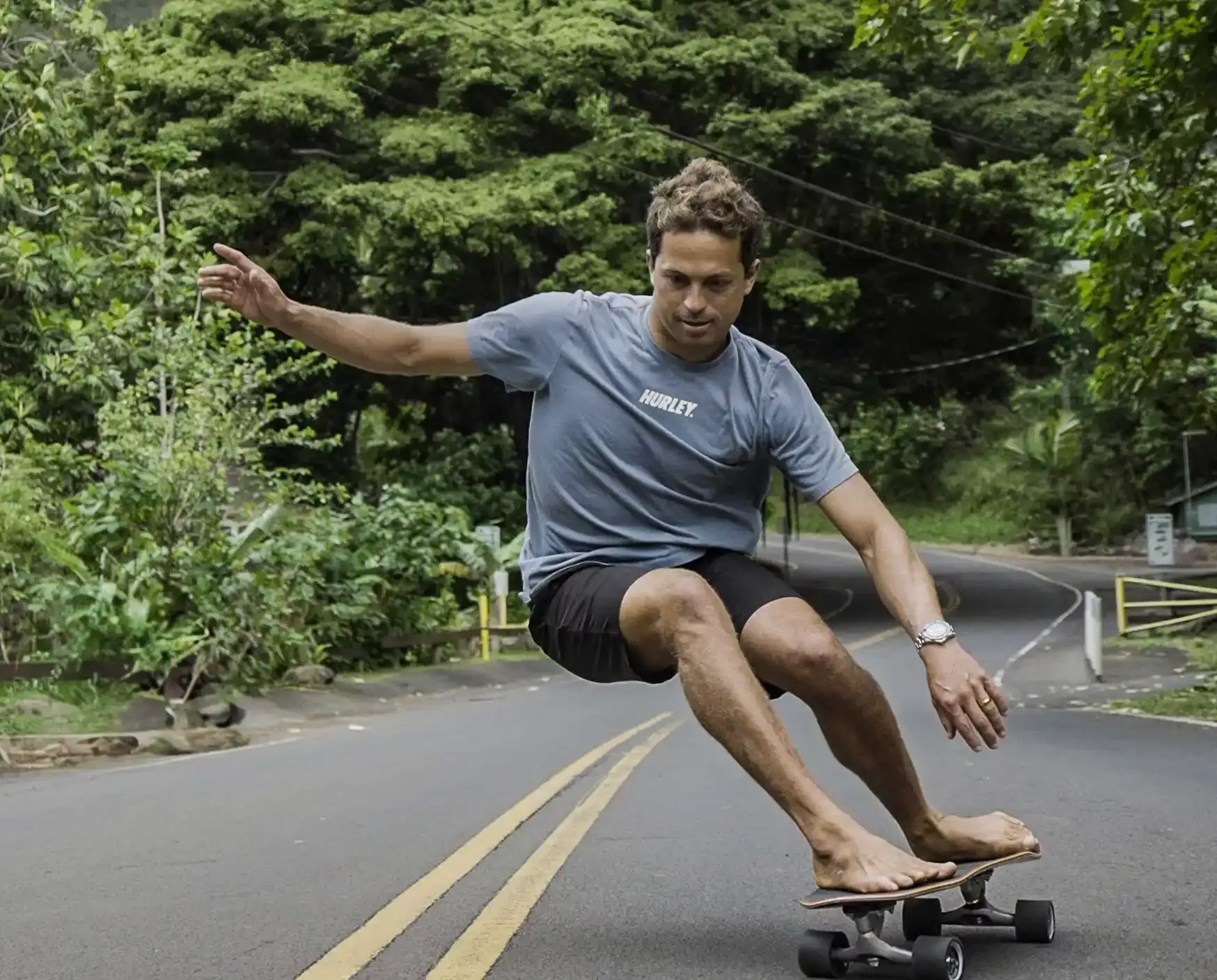 Rider su longboard scorre su strada deserta tra alberi, corpo leggermente piegato, braccia in equilibrio e look casual con maglietta grigia.