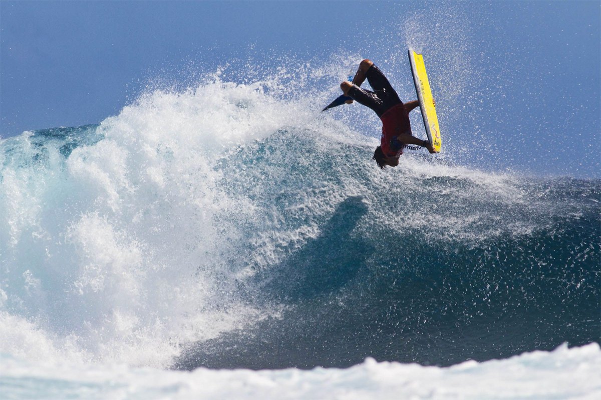 Surfer beim Bodyboard- oder Surf-Salto über einer großen Welle mit gelbem Board in klarer Meeresbrise.