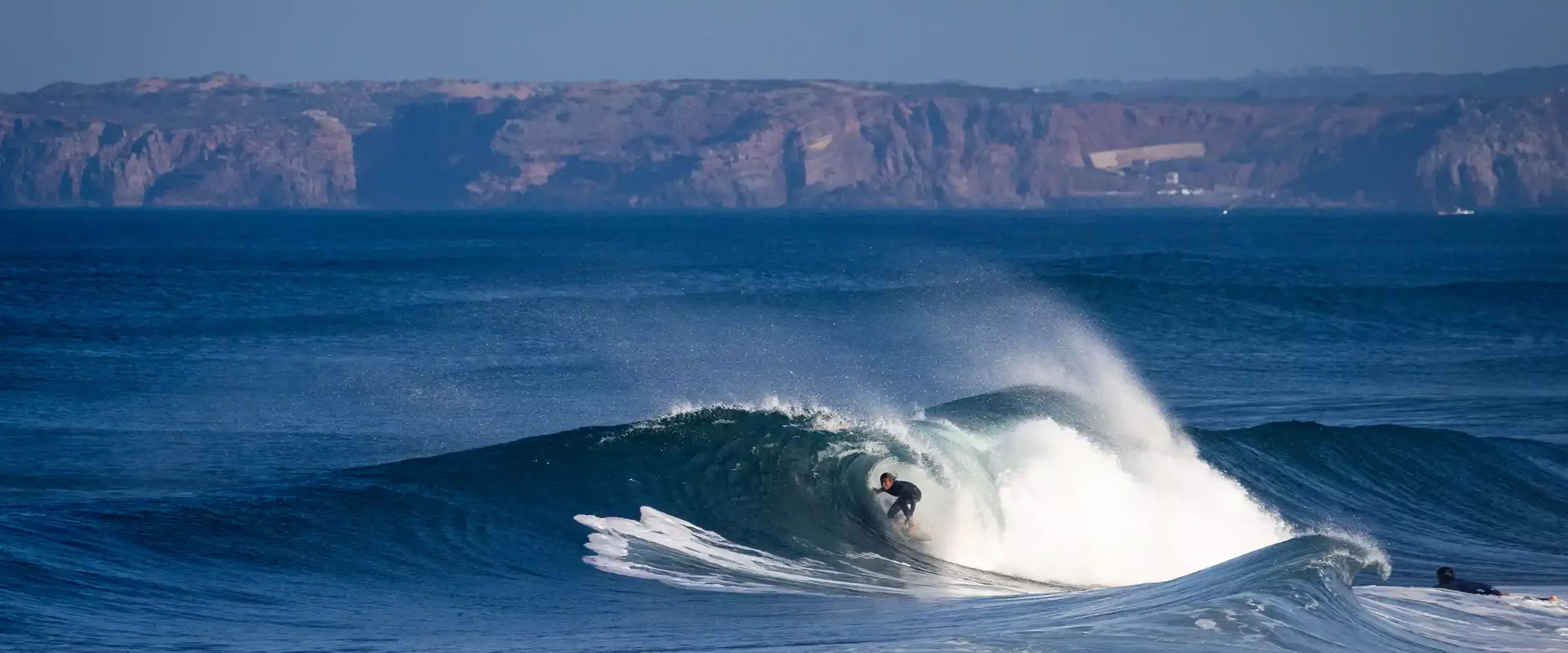 Surfer sur une vague creuse, debout sur la planche, en pleine houle bleue près de la côte rocheuse au loin.