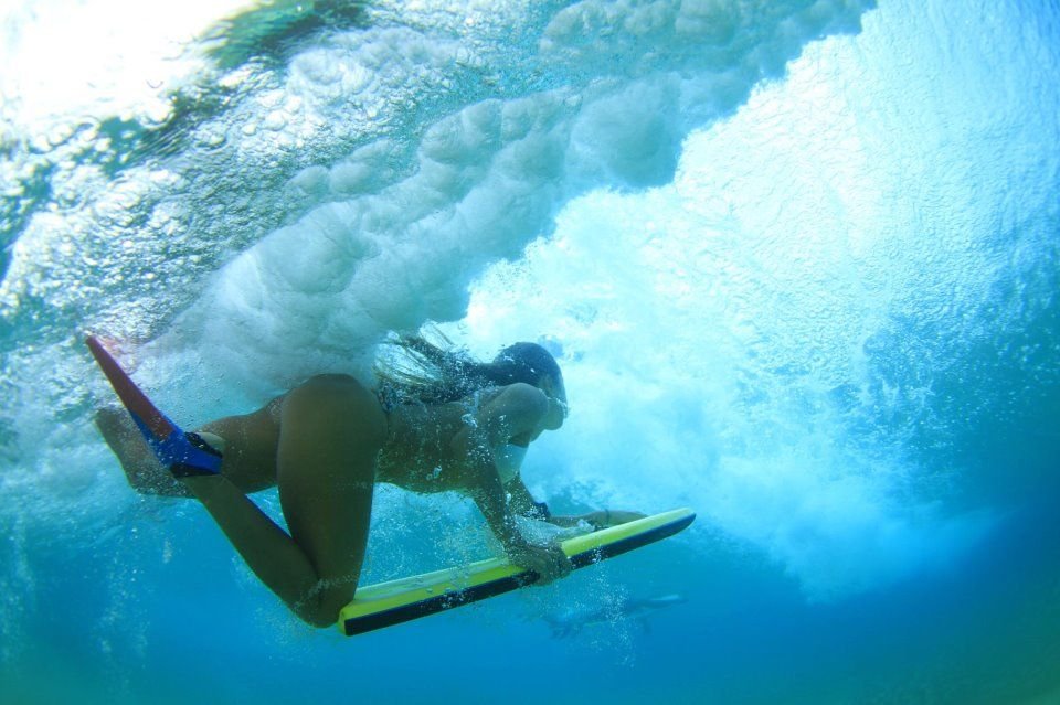 Surferin unter Wasser auf einem Bodyboard, taucht durch eine Welle mit türkisblauem Wasser.