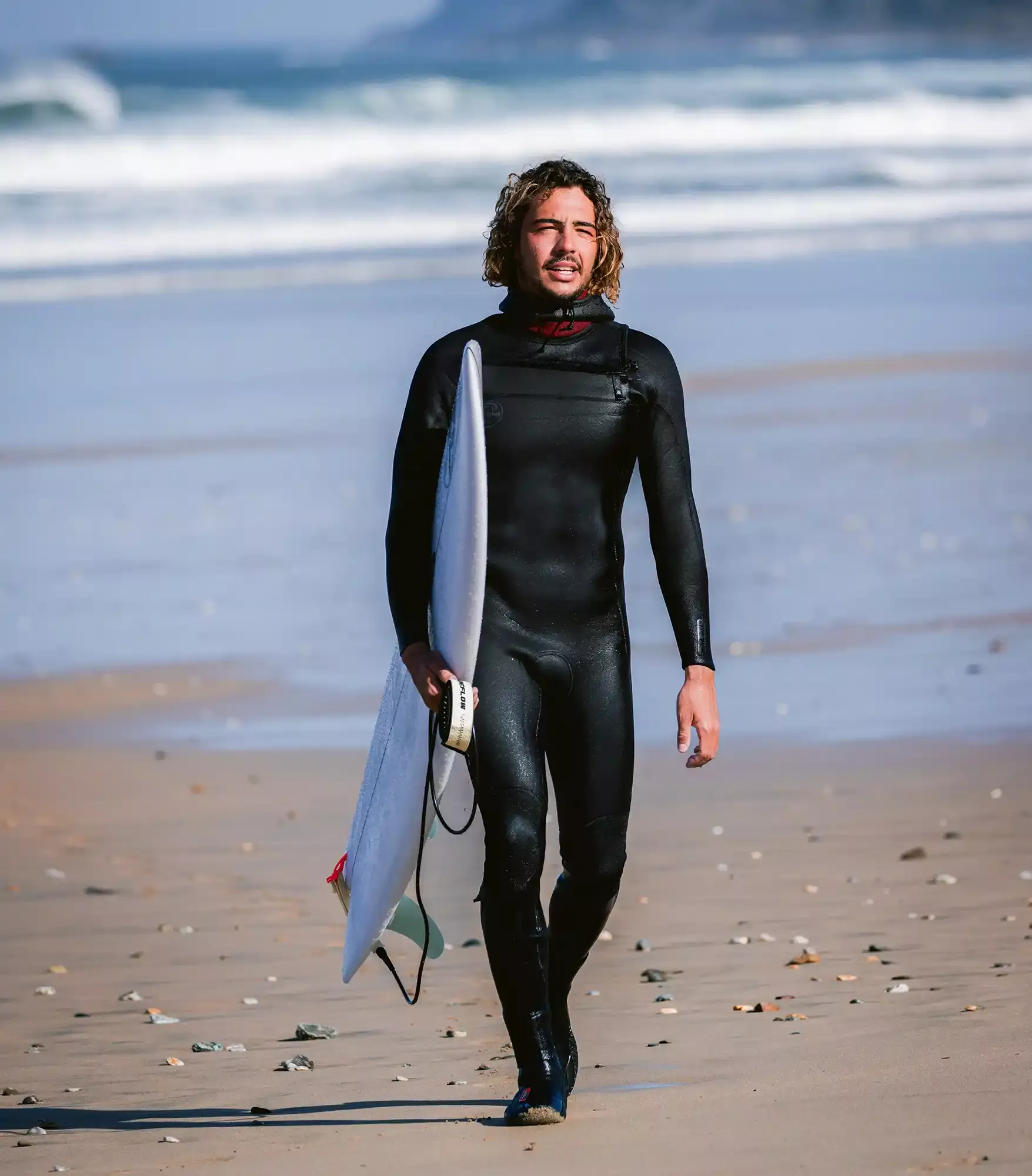 Surfeur en combinaison noire marche sur la plage avec une planche sous le bras, près des vagues bleues.