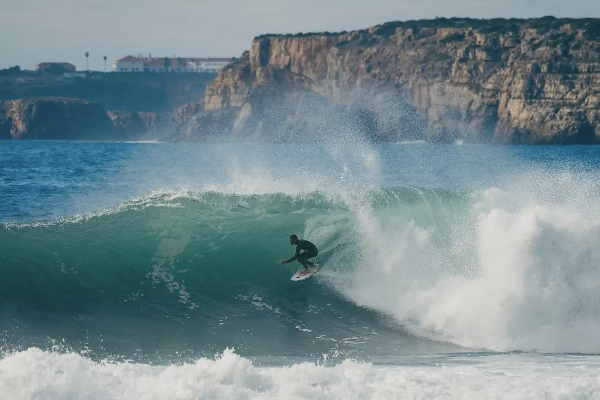 Surfista cabalgando una gran ola verde en la playa, con rocas y acantilados al fondo y agua espumosa en primer plano.
