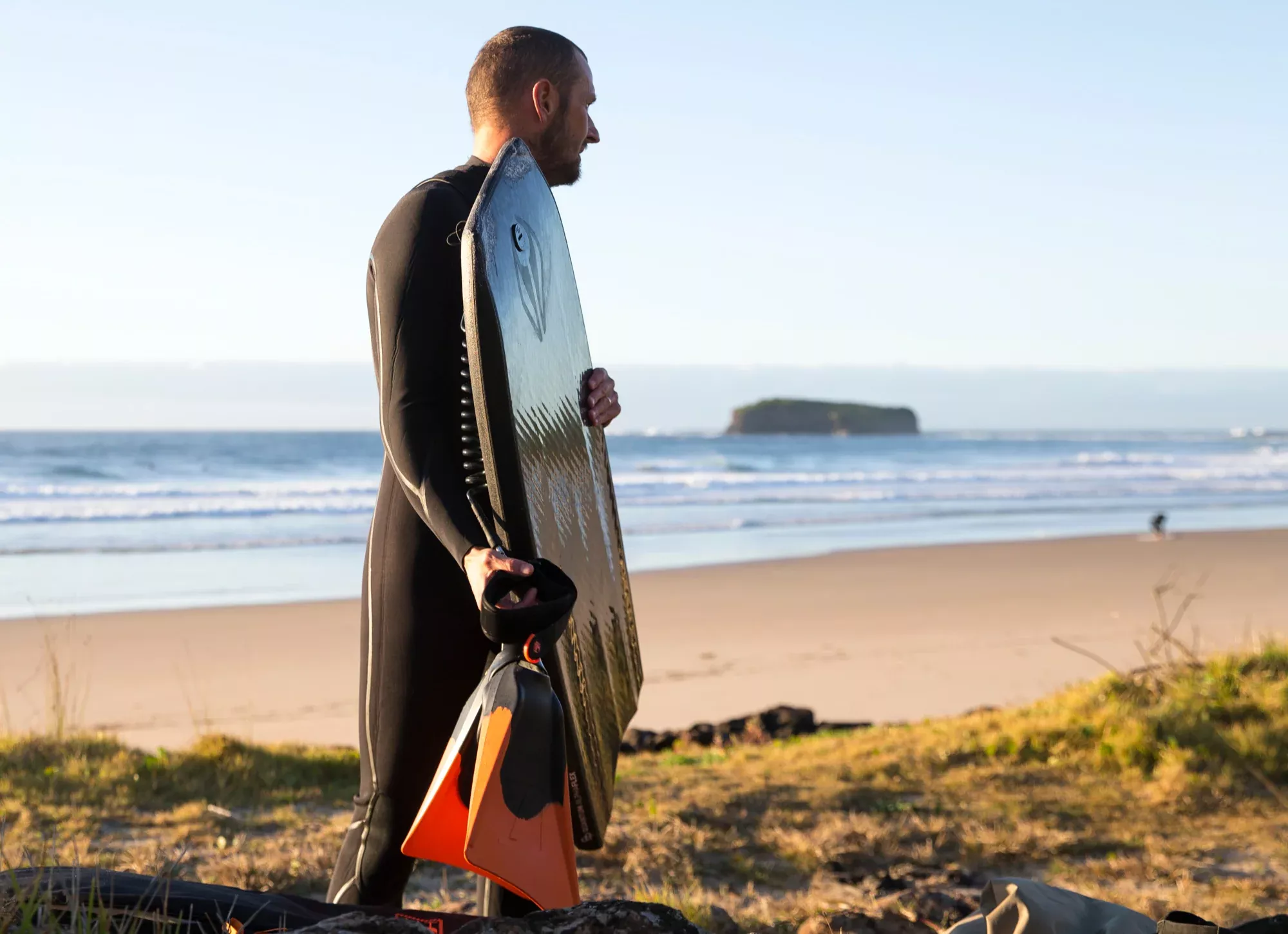 Surfista in muta sorregge tavola da surf e pinne arancioni sulla spiaggia, con mare e scoglio in lontananza.