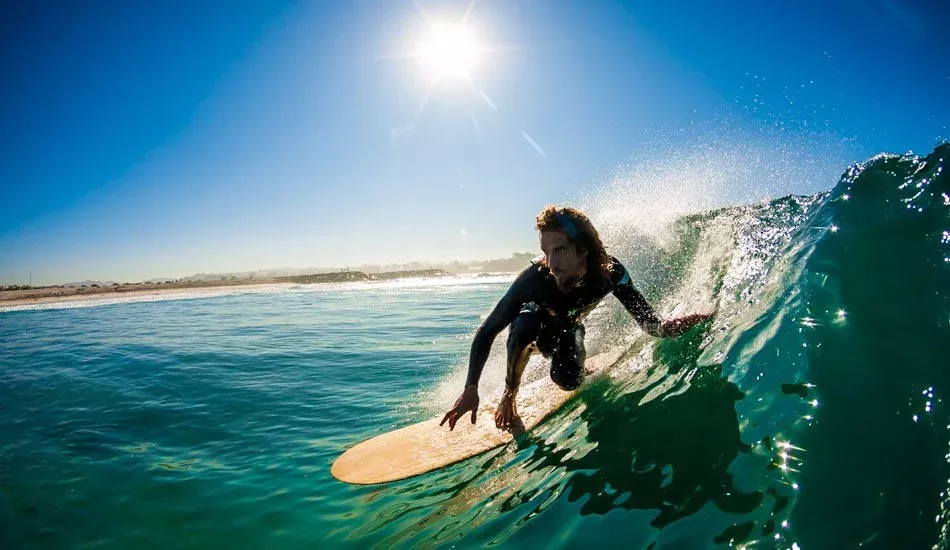 Surfista su una tavola da surf cavalca un’onda verde al largo, sole brillante in cielo azzurro.