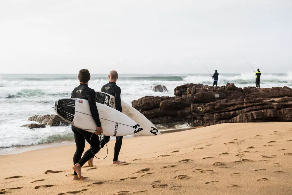 Surfistas con tablas caminan por la playa hacia las rocas para tomar ola; se ve el mar y otros pescadores al fondo.
