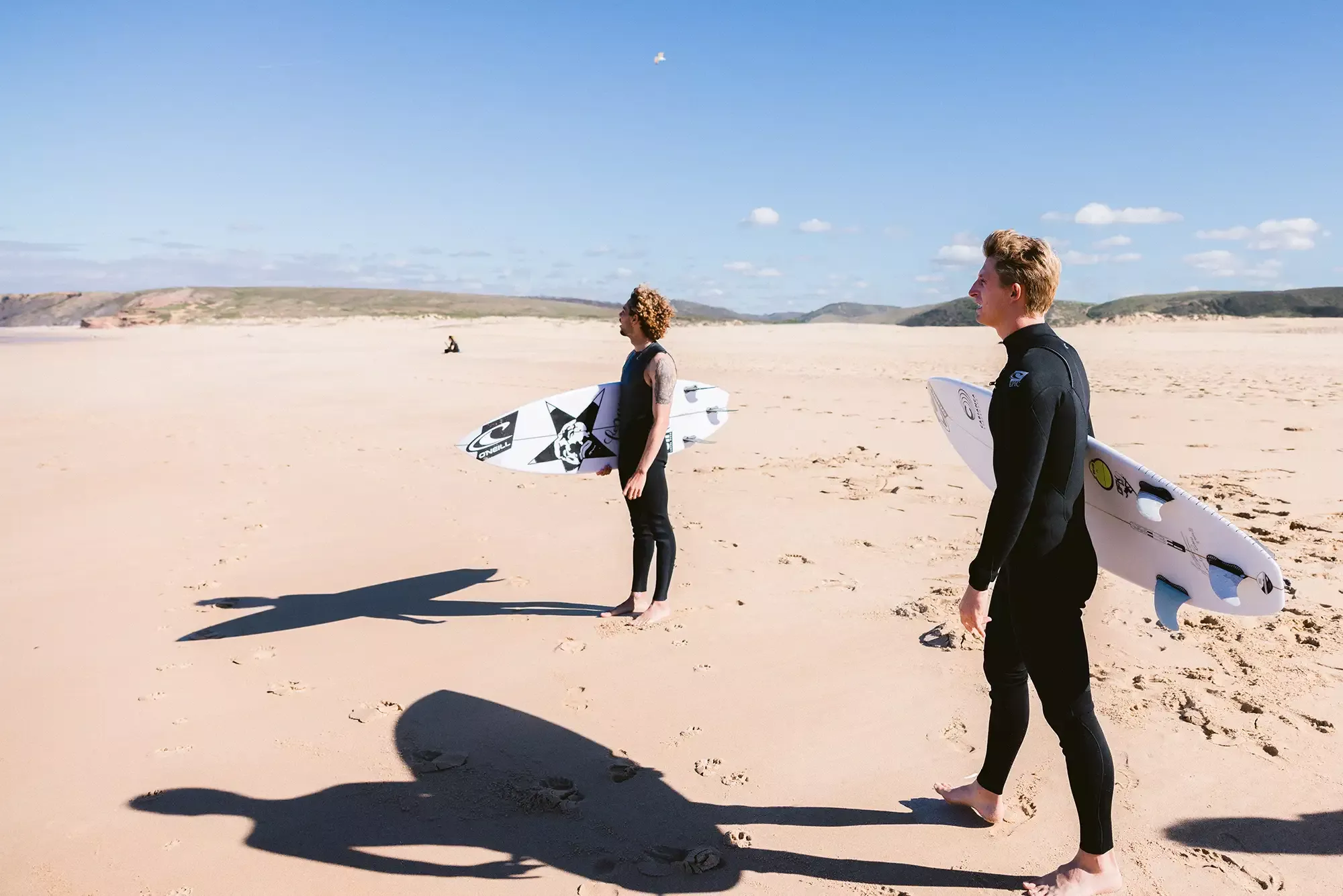Zwei Surfer am Strand mit Surfbrettern: ein Mann und eine Frau in Neoprenanzügen stehen im Sand, jeder mit einem Surfbrett unter dem Arm; lange Schatten erstrecken sich auf den Boden, strahlend blauer Himmel.
